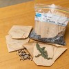 A clear zip-lock bag rests on a wooden surface. It is filled with folded brown paper packages sitting on top of small brown and white spheres of silica gel. Next to the bag are three folded packets and one unfolded packet, with dried green leaves sitting inside.