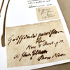 Several labels are affixed to the corner of a white piece of card. The largest is pale brown and mottled, with cursive handwriting in ink which has bled into the paper. Next to the label are two pressed, brown stems of a specimen. 
