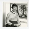 A black and white photograph of Doris Sinkora sitting by a painting of a horseback rider amongst several large, old Eucalyptus trees. Doris is smiling and holds a pair of glasses in her hand atop her lap.
